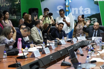 Varias personas sentadas alrededor de una mesa de reuniones con micrófonos, documentos y letreros con el logotipo del partido UNE