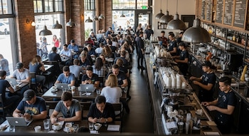 Vista aérea del interior de una cafetería moderna y concurrida en Los Ángeles, con muchos clientes sentados en mesas usando laptops y móviles, y baristas trabajando tras el mostrador.