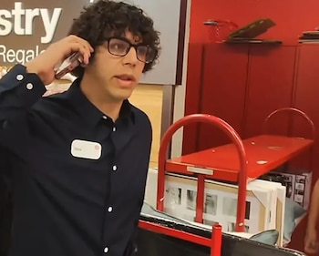 Hombre de cabello rizado y gafas habla por teléfono con una credencial en su camisa en el interior de una tienda con carros rojos
