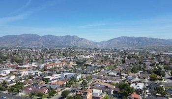 Vista aérea de un área residencial con casas de techos diversos, calles arboladas y vehículos estacionados, y montañas en el horizonte bajo un cielo azul