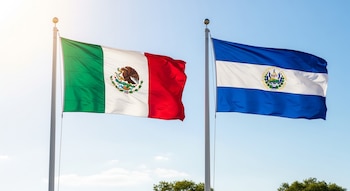 Una bandera de México verde, blanca y roja con escudo central y una bandera de El Salvador azul y blanca con escudo, ondean en mástiles bajo cielo azul.