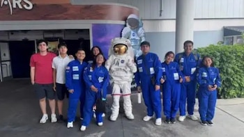 Grupo de jóvenes y adultos, algunos con overoles azules, posa con una figura de astronauta en un centro espacial. Letrero 'MARS' visible al fondo