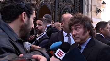 Javier Milei viste traje y corbata frente a varios micrófonos, rodeado de fotógrafos, periodistas y custodios en la entrada de un edificio.