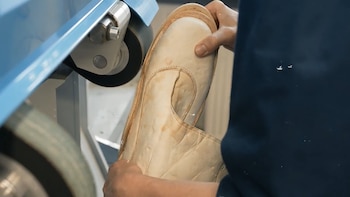 Manos de una persona sosteniendo un zapato de color crema, sin suela, siendo pulido por una máquina azul y gris con una rueda giratoria