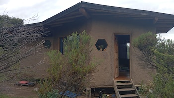 Casa rústica con paredes de barro, ventanas hexagonales y rectangulares, puerta de madera abierta y escaleras. Está rodeada de vegetación seca y verde, bajo un cielo nublado