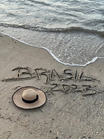 La palabra 'BRASIL 2023' escrita en la arena de una playa con la orilla del mar de fondo y un sombrero de paja con cinta negra en primer plano