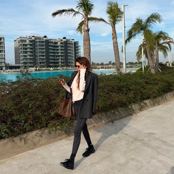 Mujer con chaqueta oscura y gafas de sol camina por un sendero junto a palmeras, laguna y edificios, sosteniendo un teléfono móvil