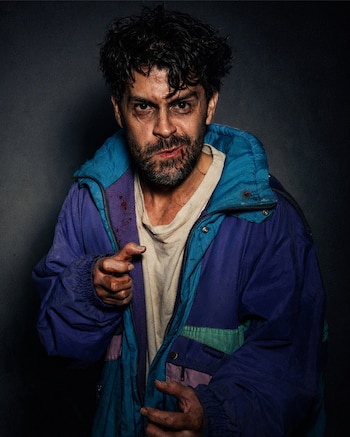 Primer plano de un hombre con barba desaliñada, rostro sucio y sangre en la boca, vestido con chaqueta azul, morada y verde, señalando al frente