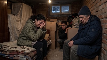 Tres personas permanecen sentadas en un sótano de ladrillos junto a cajas de cartón, mantas y una ventana nevada.