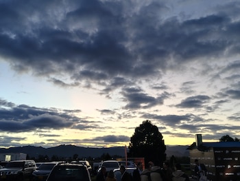 Grupo de personas junto a autos estacionados y un árbol, con montañas de fondo bajo un cielo de atardecer