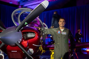 Un piloto sonriente con mono de vuelo gris se apoya en un avión rojo brillante con hélice negra de dos palas; el avión tiene la cabina abierta y luces de escenario detrás