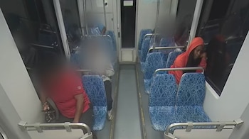 Interior de un vagón de tren con asientos azules vacíos y algunas figuras borrosas. Una persona con sudadera roja mira por la ventana