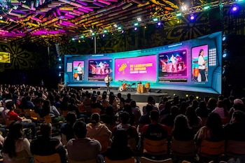 Una audiencia numerosa sentada de espaldas observa a una mujer en el escenario con una gran pantalla curva y una batería, en una feria vocacional