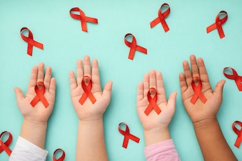 Cuatro manos de niños, de diferentes tonalidades de piel, sostienen listones rojos del VIH sobre un fondo azul claro con listones rojos dispersos.