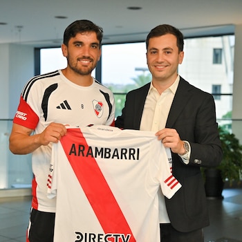Dos hombres sonrientes de pie sostienen una camiseta de fútbol blanca con banda roja y el nombre ARAMBARRI. El de la izquierda lleva una camiseta de River Plate