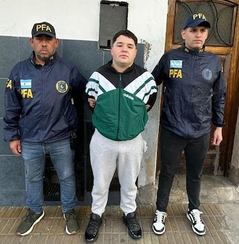 Un joven escoltado por dos efectivos con chaquetas y gorras de la Policía Federal Argentina frente a una puerta