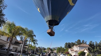 Globo aerostático azul con estrellas y lunas amarillas desciende sobre una calle residencial. Se ve su cesta de mimbre, casas, palmeras y personas en la acera