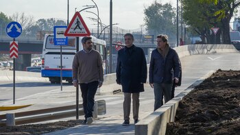 Tres hombres caminan por la vereda de una calle recién pavimentada junto a señales de tránsito, un colectivo y barreras de hormigón