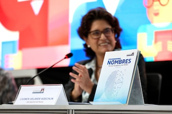 Una mujer, Carmen Velarde Koechlin, sentada frente a un micrófono y un atril con la revista Nombres, en un evento con fondo difuso