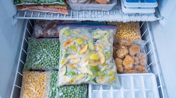 Un congelador lleno de bolsas de plástico con trozos de manzana, guisantes, maíz, alimentos empanados y cubiteras de hielo.