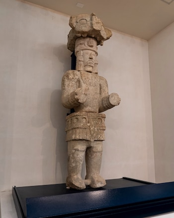 Estatua de piedra alta de figura humana con tocado en la cabeza y ornamentos, sobre un pedestal azul oscuro, contra una pared beige clara