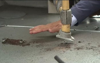 Una mano toca un poste amarillo corroído con base metálica en un piso metálico sucio con residuos de óxido, dentro de lo que parece ser un bus