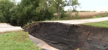 Una carretera de asfalto dividida por un gran socavón en la tierra, con tuberías expuestas, árboles y campos de maíz