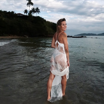 Noelia Marzol, de espaldas y mirando a cámara, parada en el agua de una playa, con un vestido de encaje blanco mojado. Fondo de colina y palmeras