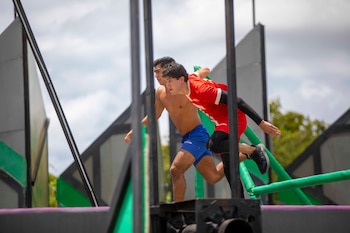 Dos atletas masculinos corren en un circuito de obstáculos al aire libre bajo un cielo nublado, con estructuras grises y verdes de fondo
