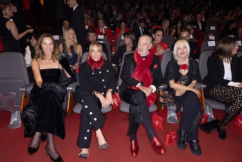 Múltiples personas, vestidas de gala, sentadas en butacas de un auditorio con suelo de moqueta roja, atentas al escenario. Se observan trajes negros y detalles rojos