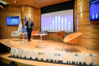 Un hombre en traje habla en un escenario con sillas, pantallas con un gráfico de barras de YPF y la inscripción #MODOCOMPLIANCE