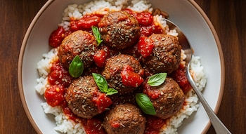 Plato hondo con arroz blanco y albóndigas en salsa de tomate, decoradas con hojas de albahaca y orégano. Se ve una cuchara metálica en el borde derecho.