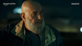 Primer plano de un hombre calvo de barba blanca con una chaqueta azul y cuello beige, mirando hacia la derecha en un ambiente oscuro