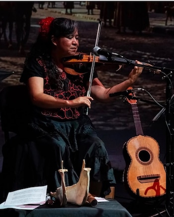 Mujer sentada que toca el violín frente a un micrófono, con una pequeña guitarra al lado y una quijada con partituras sobre una mesa