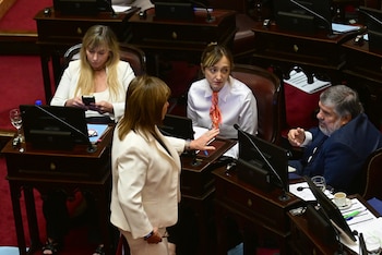 Vista aérea de cuatro legisladores en un recinto parlamentario con escritorios de madera oscura y alfombra roja, interactuando durante una sesión