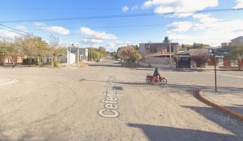 Vista urbana de una intersección de calles de tierra con casas, árboles y una persona en motocicleta