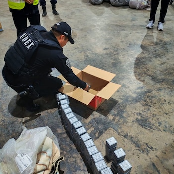Un oficial de seguridad con uniforme negro y chaleco táctico inspecciona pequeñas cajas metálicas apiladas en el suelo y dentro de una caja de cartón