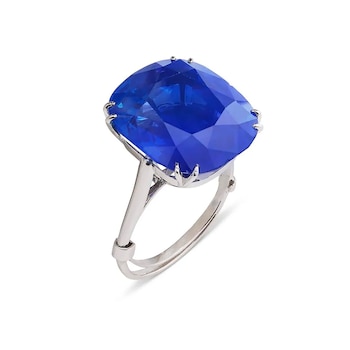 Anillo de metal plateado con un gran zafiro azul facetado de corte cojín, mostrando sus múltiples caras brillantes sobre un fondo blanco