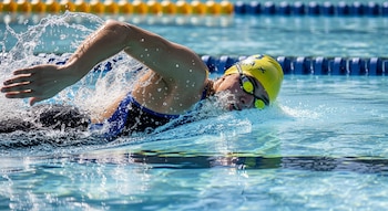 Primer plano de un nadador en una piscina, llevando gorro amarillo y gafas, mientras avanza en el agua con grandes salpicaduras alrededor de su cabeza y brazo.