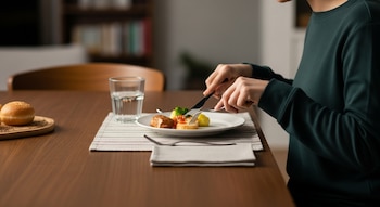 Foto horizontal de una persona sentada a una mesa de madera, cortando comida de un plato pequeño con cubiertos, con un vaso de agua y un panecillo.