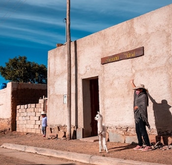 Una mujer con poncho y sombrero, una cría de alpaca en la entrada y una niña junto a un muro frente a una casa de adobe