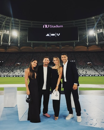 Cuatro personas vestidas elegantemente, dos hombres y dos mujeres, posando en un estadio de noche con una gran pantalla mostrando el logo "nu Stadium"
