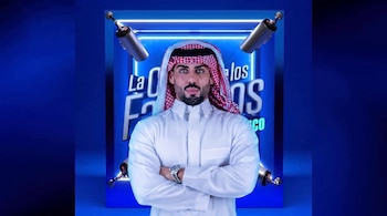 Hombre con ghutra y thobe blanco, brazos cruzados, frente a un fondo azul con texto "La Casa de los Famosos México" y dos micrófonos.