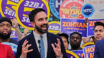 Homem barbudo, Zohran Mamdani, de terno azul e gravata, falando ao microfone aberto, de boca aberta, cercado por pessoas segurando sinais de apoio