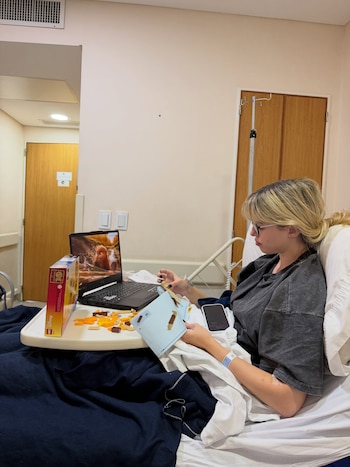 Mujer rubia en cama de clínica con vía intravenosa, armando un set de construcción Lego sobre una mesa con un manual, piezas, un portátil y un móvil