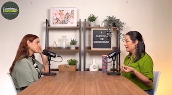 Dos mujeres sentadas frente a frente en una mesa de madera con micrófonos. Detrás de ellas, estantes con plantas y un letrero que dice "Cuestión de Cuestionar"