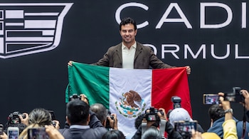Sergio Pérez sonríe mientras sostiene una bandera de México frente a una pantalla con la palabra Cadillac y varias cámaras fotografían la escena.