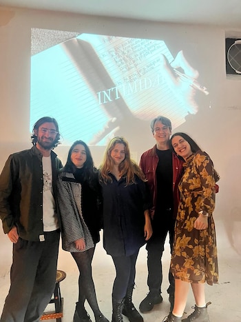 Cinco personas de pie posan sonrientes frente a una pared blanca con la proyección de un cuaderno manuscrito