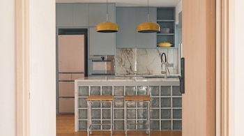 Vista de una cocina moderna con gabinetes celestes, encimera de mármol, isla con bloques de vidrio y dos taburetes. Se observa una nevera rosa y lámparas amarillas.