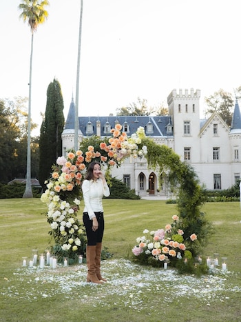 Brenda Di Aloy de pie en un jardín con un gran arco floral y velas alrededor, frente a un castillo blanco. Viste blusa blanca, pantalón negro y botas marrones